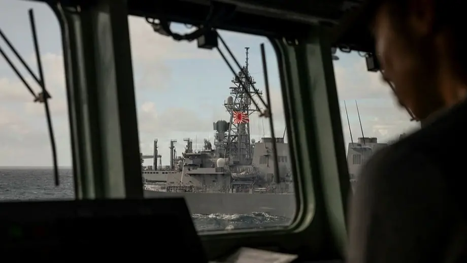 Image taken of a person in a ship cabin. Through the forward windows, in focus is a large ship bearing the Ensign of the Japan Maritime Self-Defence Force.